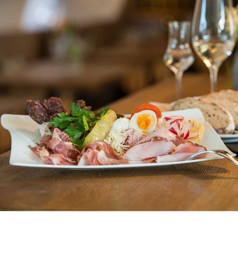 Ein Teller voller Köstlichkeiten aus der Region. Speck, Wurst, Käse, Kenn, Eier u.v.m. Im Hintergrund ein Wein- und Schnapsglas mit einem Teller voll Brotscheiben