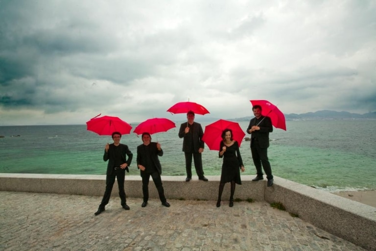 Die Bandmitglieder von Austrian Baroque stehen mit Regenschirmen auf der Mauer vor dem Meer