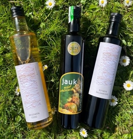 A bootle fo white wine vinegar, red wine balsam vinegar and Jauk pumpkin seed oil are lying on a green meadow