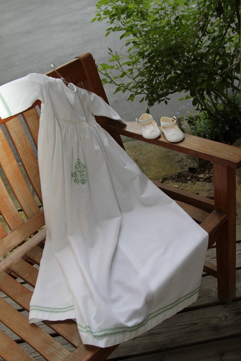 White christening gown lying on a wooden bench with little white shoes in the outdoor area of Seggau Castle.