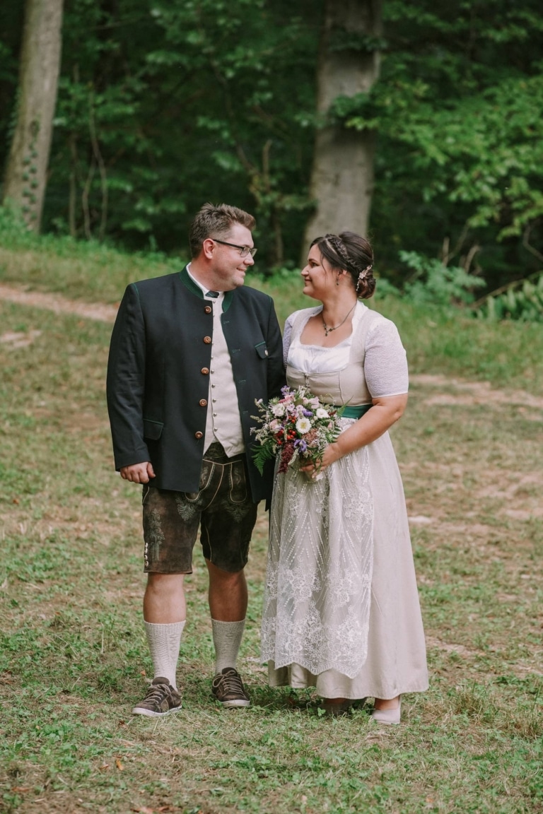 Brautpaar für die Waldhochzeit beim Schloss Seggau steht vor dem Wald, gekleidet in Tracht und sieht sich in die Augen.