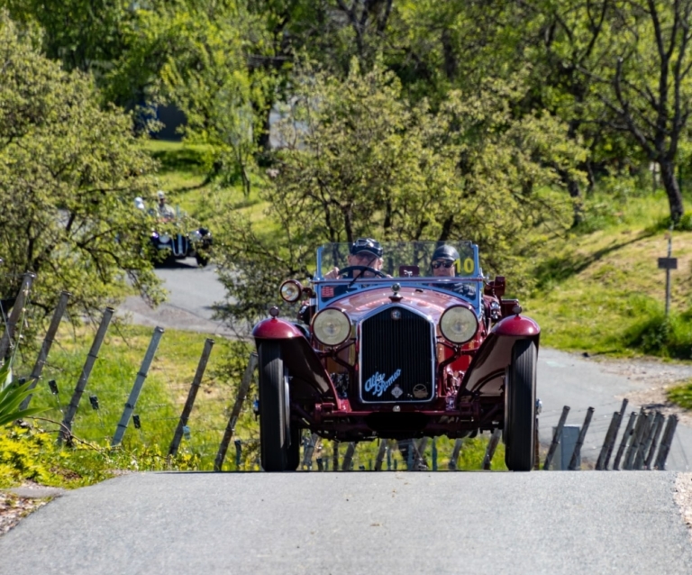 Ein Oldtimer fährt auf einer kurvigen Straße auf der Weinstraße. Im Hintergrund sind die Weinberge, ein weiterer Oldtimer und ein Klapotetz zu sehen.